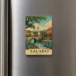 May include: A rectangular magnet featuring a scenic illustration of Salado, Texas. The image depicts a stone bridge over a river, a church, and buildings under a blue sky. The word "SALADO" and "Texas" are printed on the magnet.