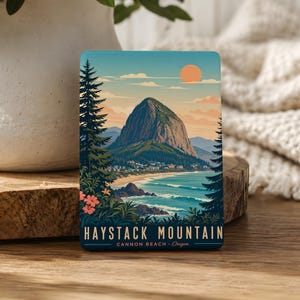 May include: A rectangular sticker featuring a scenic illustration of Haystack Mountain in Cannon Beach, Oregon. The artwork depicts a mountain, ocean, beach, and trees under a sunset sky. The text "HAYSTACK MOUNTAIN" and "CANNON BEACH - Oregon" are displayed.