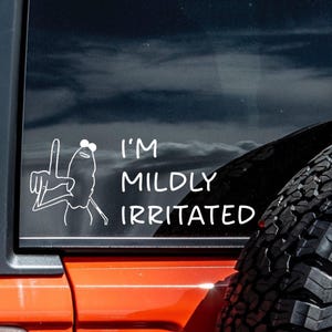 Könnte beinhalten: Weißer Vinyl-Aufkleber mit einer Cartoon-Fröschchen-Figur, die das Friedenszeichen zeigt, und dem Text "I'M MILDLY IRRITATED". Der Aufkleber befindet sich auf einem Fahrzeugfenster, mit einer orangefarbenen Karosserie und einem schwarzen Reifen.