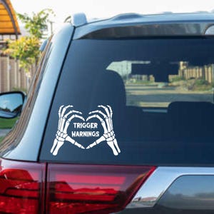 Peut inclure: Autocollant de voiture en vinyle blanc représentant un cœur formé par deux mains de squelette. Le texte "TRIGGER WARNINGS" est centré à l'intérieur du cœur. L'autocollant est appliqué sur la vitre arrière d'un véhicule.