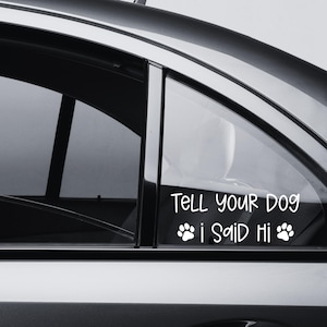 Op de afbeelding: Een witte vinyl sticker op een autoruit met de tekst "TELL YOUR DOG i SAID Hi" in een informeel, afgerond lettertype, vergezeld van twee kleine pootafdruk-pictogrammen, één aan weerszijden van de tweede regel tekst.