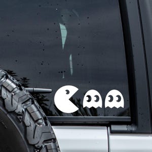 Puede incluir: Un adhesivo de vinilo blanco aplicado en la ventana oscura de un vehículo, que muestra al icónico personaje circular con la boca abierta en forma de cuña, seguido de dos figuras de fantasmas con grandes ojos redondos y bordes festoneados.