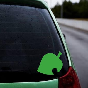 Op de afbeelding: Een felgroene sticker in de vorm van een blad, bevestigd op de achterruit van een groene auto. De sticker heeft een gestileerd bladerontwerp met een kenmerkende afgeronde inkeping aan de rechterkant. De auto staat buiten op een natte dag.