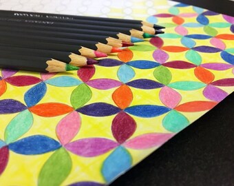 May include: A close-up of a coloring page with a repeating pattern of colorful flower shapes. A set of black colored pencils are lying on top of the page.