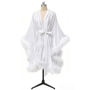 May include: A white satin robe with a V-neck, long sleeves, and a matching belt. The robe is trimmed with white feathers at the cuffs and hem. The garment is displayed on a mannequin.