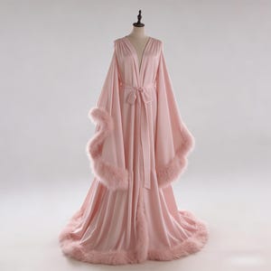 May include: A light pink satin robe with a V-neck and long, flowing sleeves. The robe features a matching belt and is trimmed with fluffy, light pink feathers along the sleeves and hemline. The robe is displayed on a mannequin.