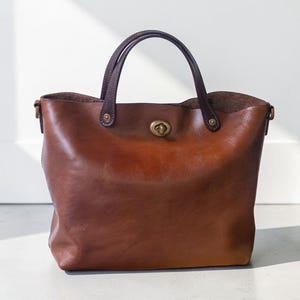 Puede incluir: Un bolso tote de cuero marrón con asa superior y cierre de broche de latón. El bolso tiene una forma estructurada con un interior espacioso. Las asas son de un marrón más oscuro.