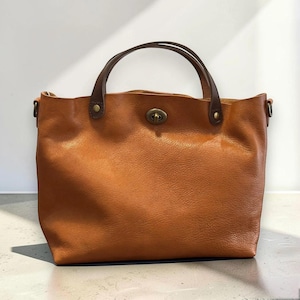 May include: A tan leather handbag featuring a smooth, textured surface, two dark brown leather handles attached with brass rivets, and a central brass turn-lock closure. The bag has a wide, open-top design with side rings for a shoulder strap.