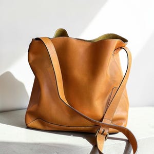 May include: A tan leather tote bag with a long shoulder strap and a buckle closure. The bag has a soft, unstructured shape and a light brown interior. The bag is sitting on a white surface.