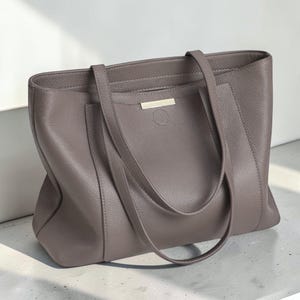May include: A taupe-colored tote bag with a pebbled texture, featuring two long shoulder straps, a rectangular silhouette, and a small horizontal metallic bar accent centered on the upper front panel above a circular stitched detail.