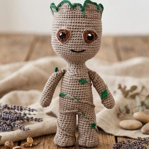 Puede incluir: Una figura de ganchillo de un personaje con apariencia de árbol, con cuerpo marrón claro, ojos marrones y detalles verdes. La muñeca hecha a mano está sobre una superficie de madera con lavanda seca y otros elementos decorativos.