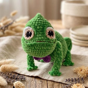 May include: A handmade green crochet chameleon with large, expressive eyes and a small purple bow tie. The plush toy is positioned on a wooden surface, showcasing its detailed craftsmanship and whimsical design. The chameleon's body is a vibrant green, with a textured, soft appearance.