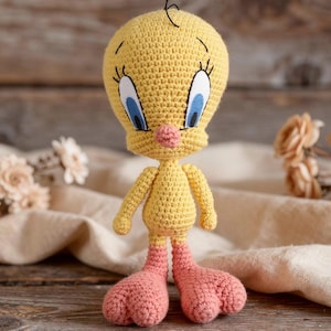 May include: A handmade crochet Tweety Bird doll. The doll is primarily yellow with a large head, blue eyes, and a pink beak. The feet are pink. The doll is standing on a wooden surface.