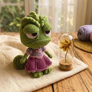 May include: A handmade green crochet chameleon doll with a purple and pink dress. Next to the doll is a yellow crochet flower in a glass dome. The doll is sitting on a white cloth.