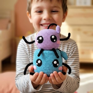 Può includere: Una coppia di peluche all'uncinetto impilati, composta da una piccola creatura color lavanda appoggiata su una creatura blu più grande di forma quadrata. Entrambe le figure hanno arti in filato nero, grandi occhi circolari bianchi e neri.