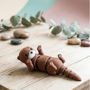Puede incluir: Una figura de nutria articulada marrón con pecho blanco y patas negras yace sobre una superficie de madera. El juguete está segmentado, lo que permite el movimiento. El fondo presenta piedras y vegetación.