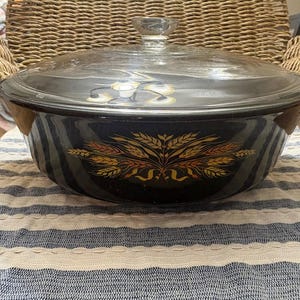 May include: A dark blue enamel casserole dish with a clear glass lid. The dish features a wheat design in gold and orange. The lid has a clear glass knob. The dish is sitting on a blue and white striped cloth.