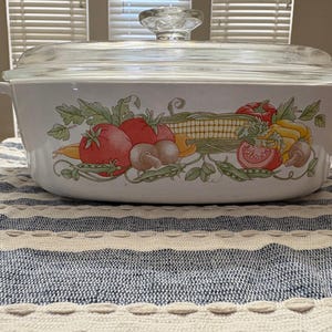 Pode incluir: Uma travessa branca com tampa de vidro transparente. A travessa é decorada com uma ilustração colorida de vegetais, incluindo tomates, milho e cogumelos. A travessa está sobre uma toalha de mesa listrada.