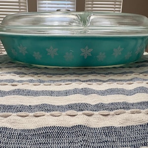 May include: A teal Pyrex casserole dish with a clear glass lid. The dish features a snowflake pattern in white. The dish is sitting on a blue and white striped tablecloth.