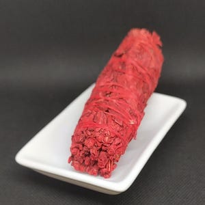 Puede incluir: Un manojo de salvia roja descansa sobre un plato de cerámica blanco sobre un fondo negro. La salvia está atada con hilo rojo, mostrando una estética vibrante y natural. Este artículo se utiliza para la limpieza espiritual y aromaterapia.