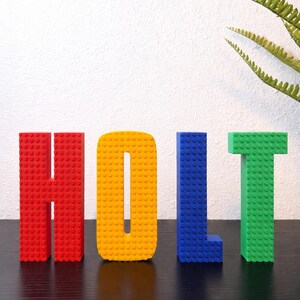 May include: A colorful display of the name "HOLT" spelled out with large, block-style letters. The letters are red, yellow, blue, and green, resembling building blocks. The letters stand on a dark surface against a white background.