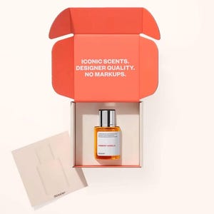 May include: An open orange box with the text "ICONIC SCENTS. DESIGNER QUALITY. NO MARKUPS." contains a bottle of "AMBERY VANILLA" perfume. A small card with a perfume bottle outline is next to the box. The perfume bottle is rectangular with a silver cap.