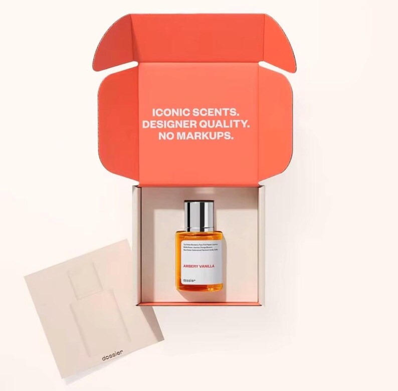 May include: An open orange box with the text "ICONIC SCENTS. DESIGNER QUALITY. NO MARKUPS." contains a bottle of "AMBERY VANILLA" perfume. A small card with a perfume bottle outline is next to the box. The perfume bottle is rectangular with a silver cap.