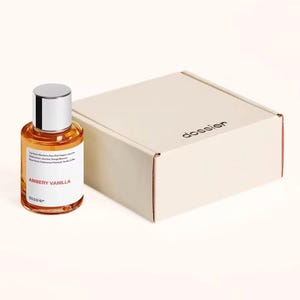 May include: A perfume bottle with a silver cap and a clear glass body filled with amber-colored liquid. The label reads "AMBERY VANILLA" and "dossier." Beside it is a beige box with the word "dossier" printed on it.