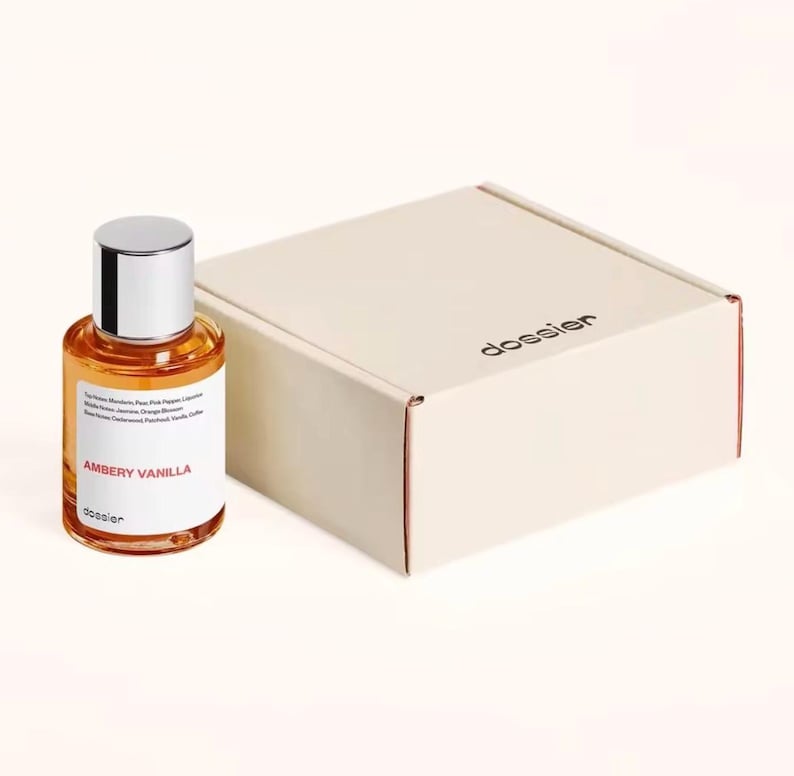 May include: A perfume bottle with a silver cap and a clear glass body filled with amber-colored liquid. The label reads "AMBERY VANILLA" and "dossier." Beside it is a beige box with the word "dossier" printed on it.