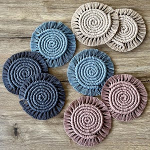 May include: A collection of handmade macrame coasters in various colors, including blue, gray, and beige. Each coaster features a spiral design with a fringed edge. The coasters are arranged on a wooden surface.