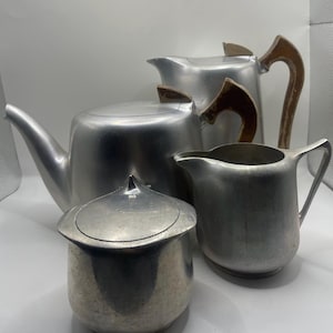 May include: A silver-toned metal tea service set consisting of a large teapot with a curved spout, a smaller lidded sugar bowl with a pointed finial, a creamer pitcher, and a tall coffee pot featuring a distinct brown wooden handle.