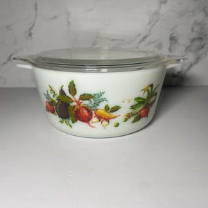 May include: White casserole dish with a clear lid. The dish features a colourful vegetable design with illustrations of beets, carrots, and other produce. The dish has two handles and is suitable for cooking and serving.