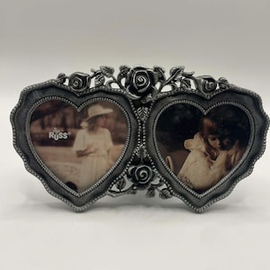 Könnte beinhalten: Ein silberfarbener Doppel-Fotorahmen mit herzförmigen Öffnungen. Der Rahmen ist mit Rosen und Blättern verziert. Die Fotos zeigen zwei Personen. Das Wort "RUSS" ist auf dem linken Foto zu sehen.