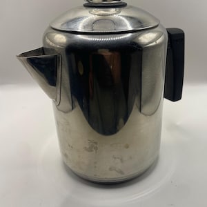 Puede incluir: Una cafetera percoladora de acero inoxidable pulido con un mango negro resistente al calor, una tapa de acero inoxidable a juego y un pomo de cristal transparente en la parte superior. La cafetera tiene un pico ancho y un cuerpo cilíndrico.