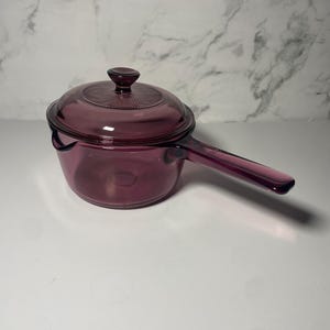 Puede incluir: Una cacerola de vidrio de color amatista con tapa. La cacerola tiene un mango largo y una tapa redonda con un pomo. El vidrio es transparente, lo que permite ver el contenido. Utensilio de cocina para usar en la estufa.