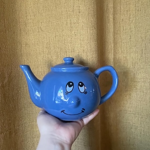 Puede incluir: Una tetera de cerámica en color azul bígaro brillante que presenta un rostro con ojos grandes mirando hacia arriba, una nariz pequeña y redondeada y una boca sonriente. Incluye una tapa a juego con un pomo y un asa lateral curva.