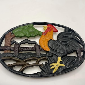 May include: An oval cast iron trivet featuring a relief design of a rooster with a red comb, orange neck, and black body. The scene includes a brown wooden fence, a green tree, and yellow feet, all framed within a black border with openwork details.