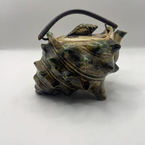 Può includere: Una teiera in ceramica a forma di grande conchiglia con una finitura lucida verde e marrone. Il coperchio presenta una piccola impugnatura a forma di conchiglia scolpita e la teiera ha un manico curvo in filo metallico marrone scuro.