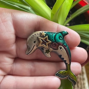 Peut inclure: Une broche en métal en forme de dauphin avec une finition argentée métallique sur la tête et la queue, rehaussée de détails en émail vert et violet vifs. Le design comprend des étoiles découpées et de petites étoiles gravées sur le corps.