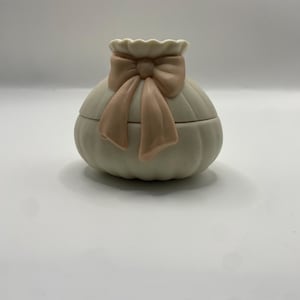 Puede incluir: Un joyero redondo de cerámica color blanco roto con una textura acanalada similar a una calabaza. La tapa presenta un lazo esculpido en color rosa claro mate centrado en la parte superior. El recipiente tiene un borde festoneado y acabado mate.