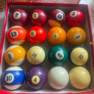 Könnte beinhalten: Ein Set aus sechzehn Billardkugeln in einer roten Schachtel. Die Sammlung enthält einfarbige und gestreifte Kugeln in verschiedenen Farben wie Rot, Orange, Gelb, Grün, Blau und Violett, die jeweils mit einer schwarzen Zahl versehen sind.