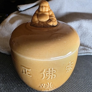May include: A tan ceramic jar with a lid featuring a small Buddha figure. The jar has Chinese characters on the front. The jar is a light brown color.