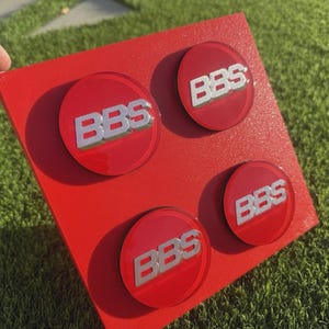 May include: A red square with four circular emblems. Each emblem has a red background with the silver letters "BBS". The items are displayed on a green surface.