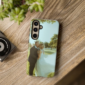 May include: A smartphone case featuring a wedding photo of a couple kissing in a park. The case has a black camera module and a light-colored border. The background is a wooden surface with a camera and greenery.