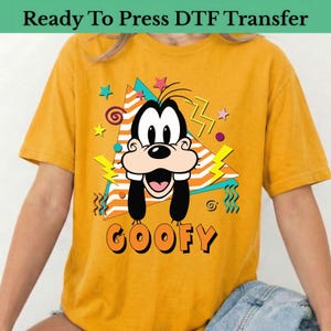 May include: Mustard yellow t-shirt featuring a graphic of Goofy with a retro design. The design includes a triangle, stars, and lightning bolts. The word "GOOFY" is printed in orange and black. The text "Ready To Press DTF Transfer" is at the top.