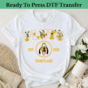 May include: White t-shirt featuring the name "PLUTO" in yellow letters with Pluto illustrations. Below, a yellow circle with Pluto's face and the text "EST. 1930 DISNEYLAND". The words "Ready To Press DTF Transfer" are at the top.