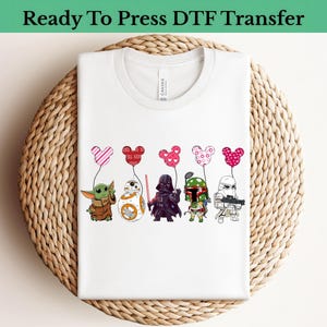 Puede incluir: Camiseta blanca con un gráfico de personajes de Star Wars sosteniendo globos con forma de Mickey Mouse. Los personajes incluyen a Baby Yoda, BB-8, Darth Vader, Boba Fett y un Stormtrooper. El texto "Ready To Press DTF Transfer" está en la parte superior.