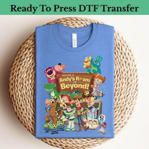 Peut inclure: Un t-shirt bleu clair avec un graphisme coloré des personnages du film "Toy Story". Le motif comprend le texte "Welcome to Andy's Room and Beyond!" et les mots "Ready To Press DTF Transfer" en haut.
