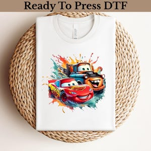 May include: White t-shirt featuring a colorful graphic of two cartoon cars, one red and one orange, with smiling faces. The design includes splashes of blue, orange, and green. The text "Ready To Press DTF" is at the top.