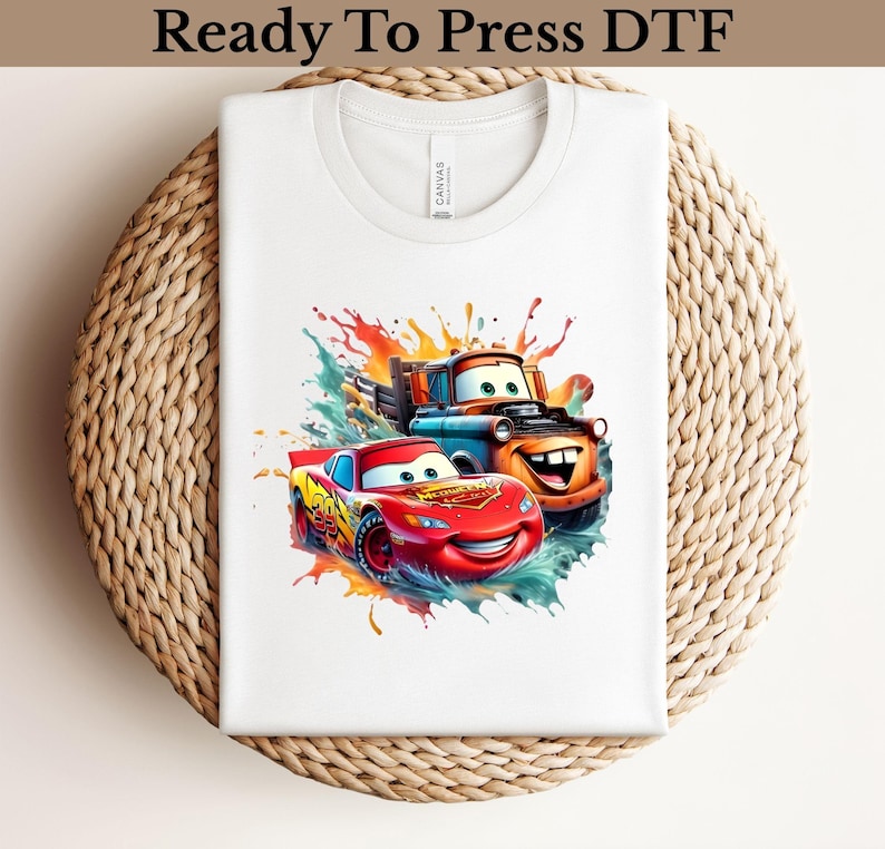 May include: White t-shirt featuring a colorful graphic of two cartoon cars, one red and one orange, with smiling faces. The design includes splashes of blue, orange, and green. The text "Ready To Press DTF" is at the top.